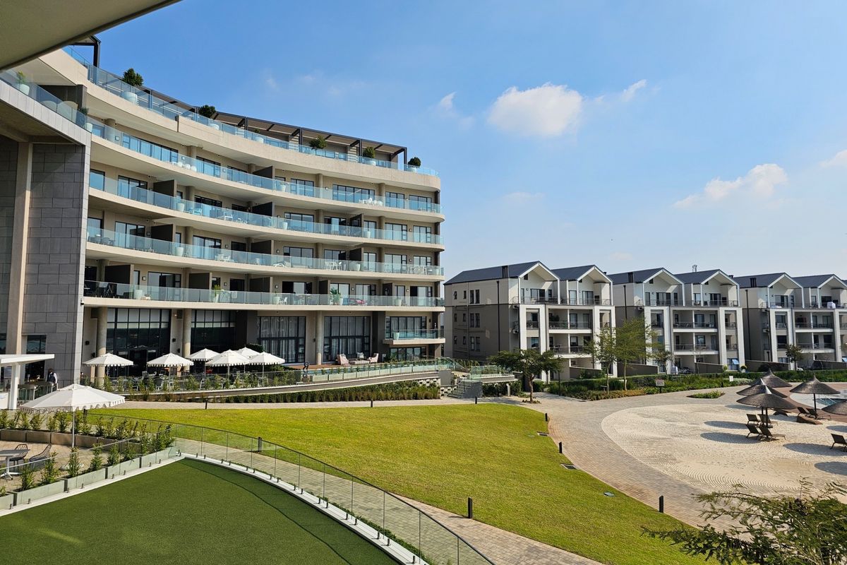 A view of the apartment complex exterior featuring modern multi-story buildings, glass balconies, landscaped gardens, and paved communal areas.