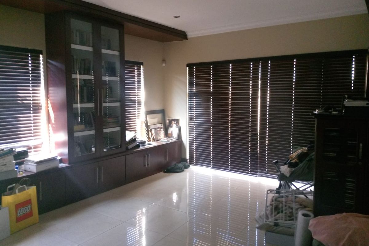 A spacious living area featuring polished tile flooring, built-in dark wood cabinetry with glass-fronted shelving, and dark horizontal window blinds.