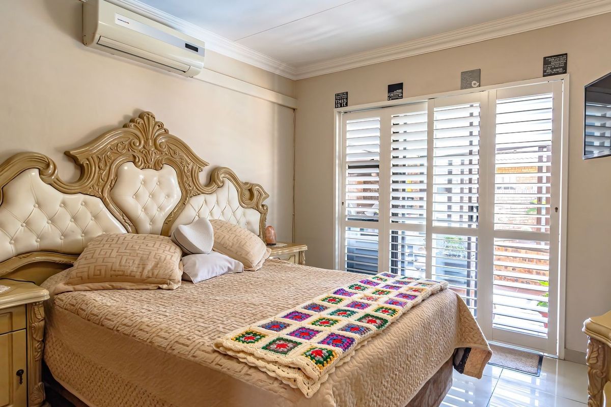 A well-maintained bedroom featuring tiled flooring, crown molding, a wall-mounted air conditioning unit, and white plantation shutters leading to an outdoor area.