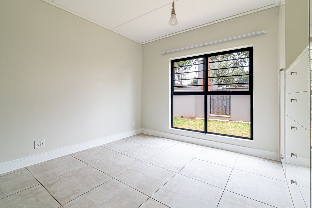 A bright, empty bedroom featuring neutral tiled flooring, built-in cupboards, and a large window with security bars overlooking the garden.