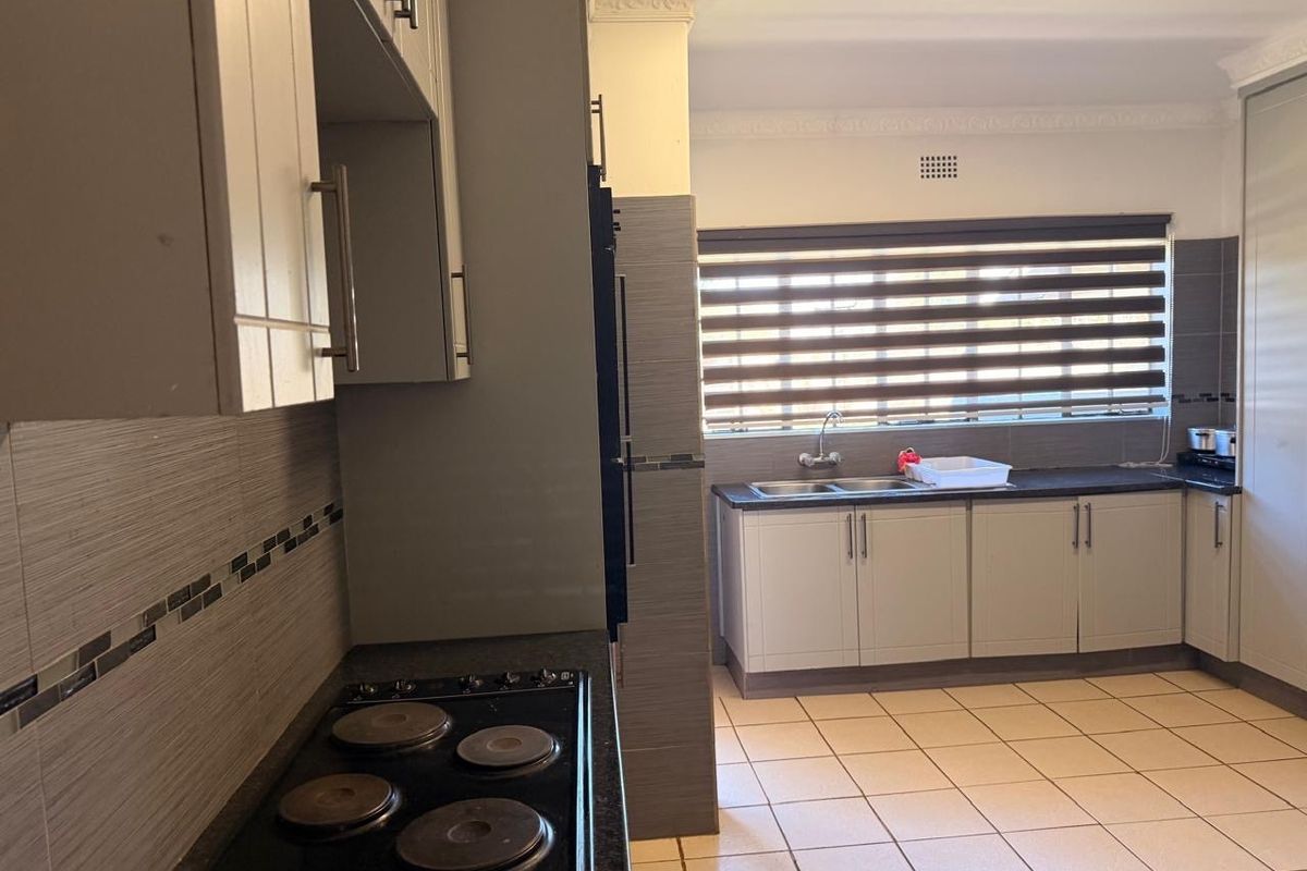 A functional kitchen featuring light-toned cabinetry, dark countertops, a built-in electric hob, and tiled flooring with a window above the sink area.