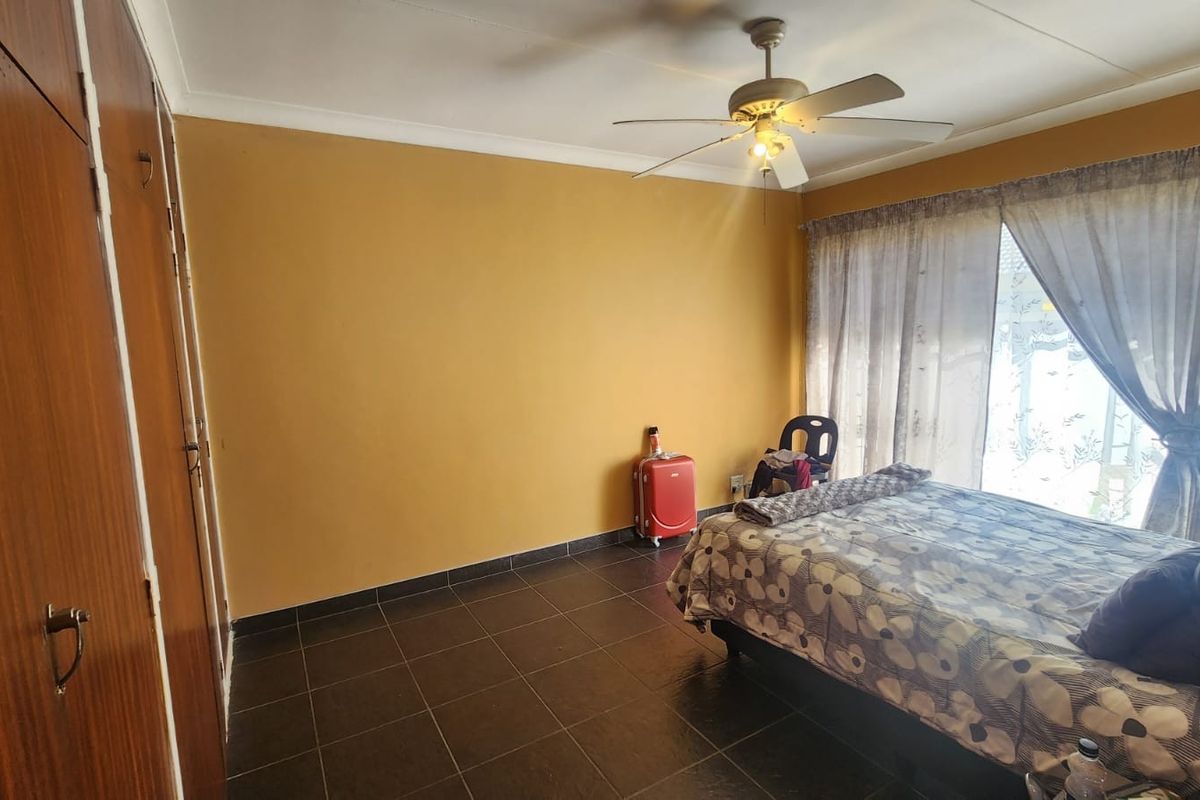 A bedroom featuring dark floor tiles, built-in wooden cupboards, and a ceiling fan, offering a functional space with natural light.