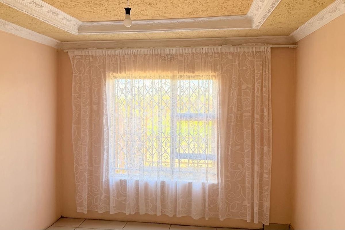 A simple bedroom featuring tiled flooring, a textured ceiling with decorative cornice, and a window fitted with security bars.