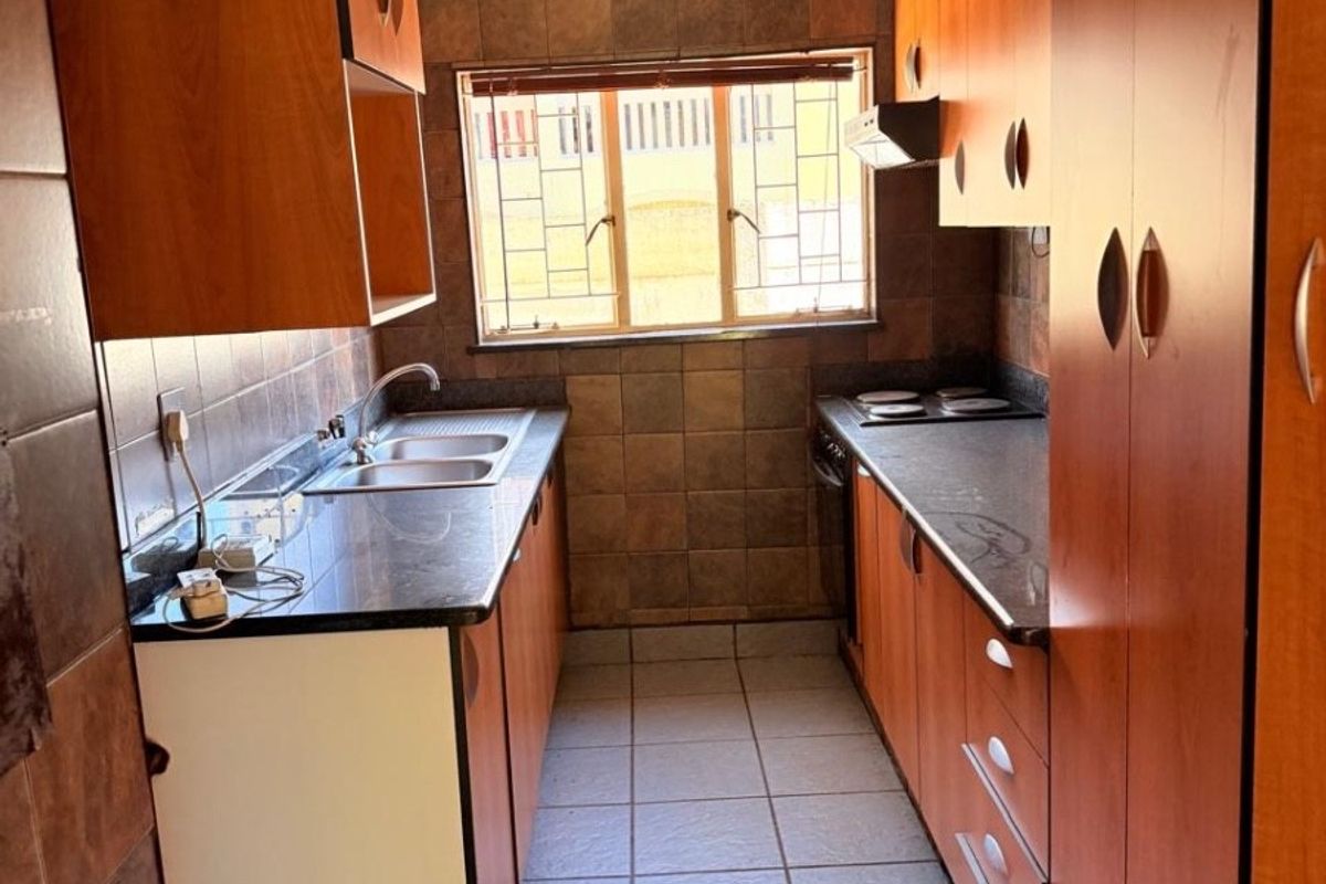A galley-style kitchen featuring wooden cabinetry, dark granite countertops, a built-in hob, and tiled flooring with a window providing natural light.