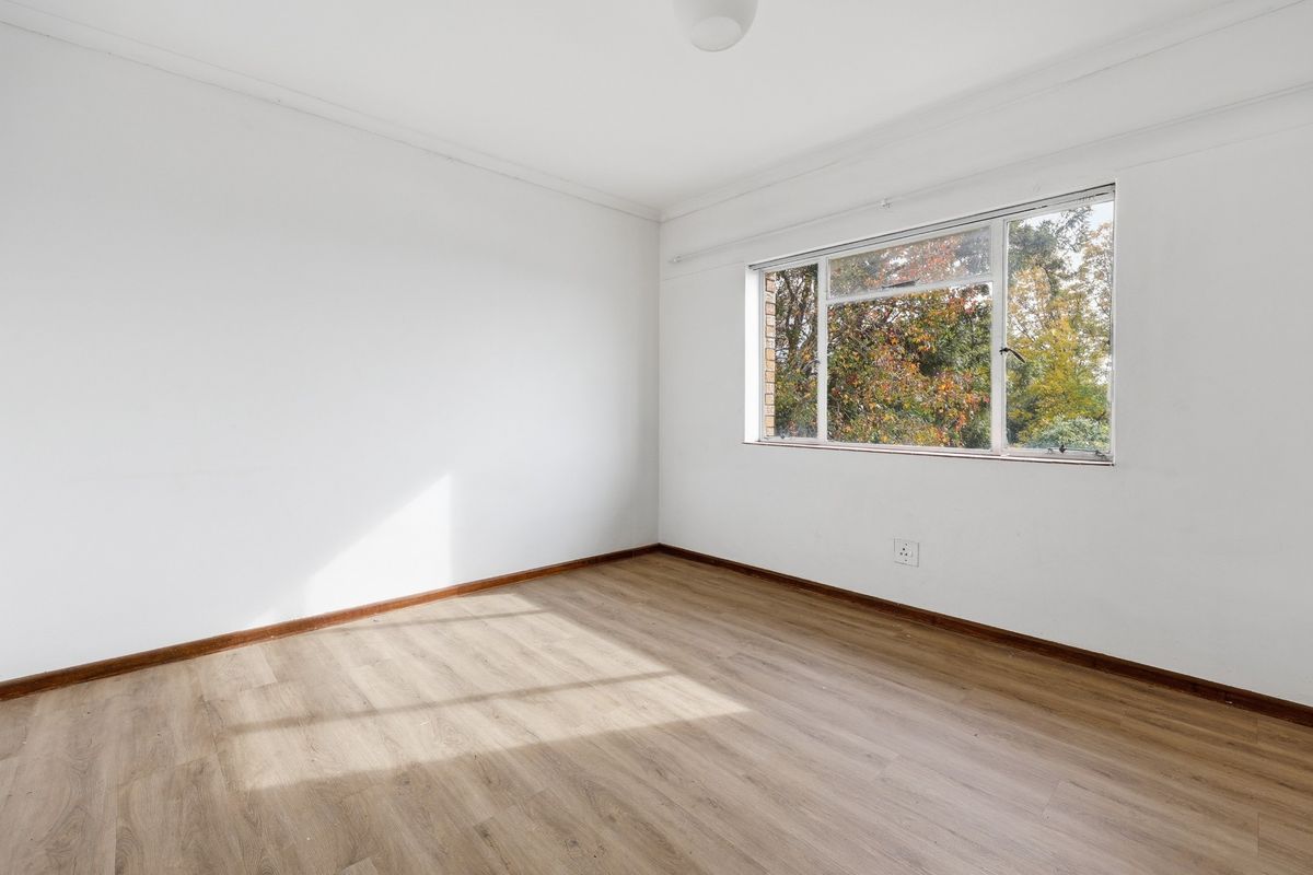 A bright, empty bedroom featuring light wood-look flooring, white walls, and a large window overlooking mature trees.
