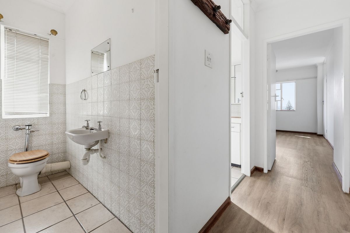 A functional bathroom featuring a wall-mounted basin and toilet, with tiled walls and wood-look flooring, leading into an adjacent living area.