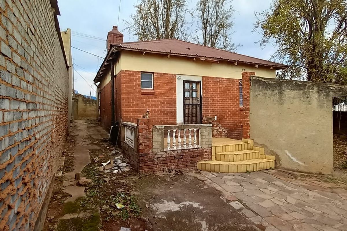 Rear view of a brick house featuring a small tiled entrance, concrete balustrade, and surrounding boundary walls in need of maintenance.