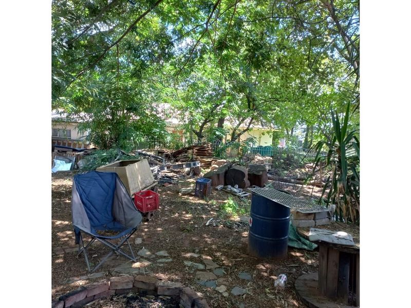 A large, shaded garden area featuring mature trees and a brick-lined fire pit, currently requiring significant clearing and landscaping to restore its potential.