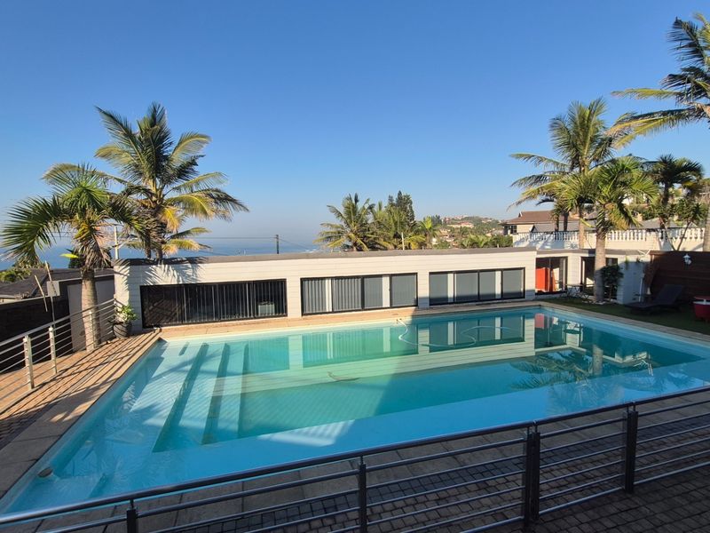 A spacious outdoor pool area featuring a large swimming pool, paved surround, stainless steel safety railing, and established palm trees.