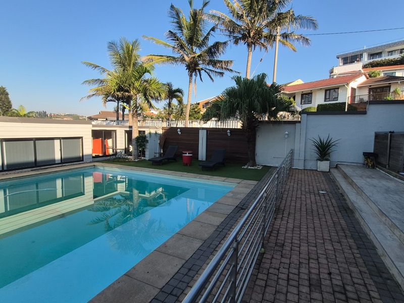A well-maintained outdoor pool area featuring a paved deck, stainless steel safety railing, and landscaped garden space with palm trees.