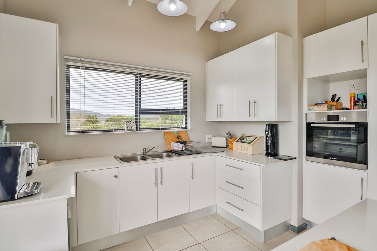 A bright, modern kitchen featuring white cabinetry, stone-look countertops, a double stainless steel sink, and a built-in oven with a view of the outdoors.