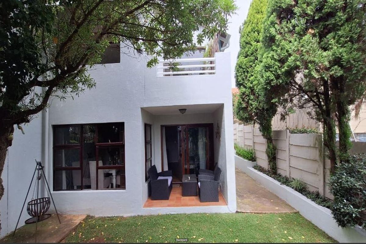 Rear view of the townhouse featuring a covered patio, small garden, and an upper-level balcony with established trees and perimeter walling.