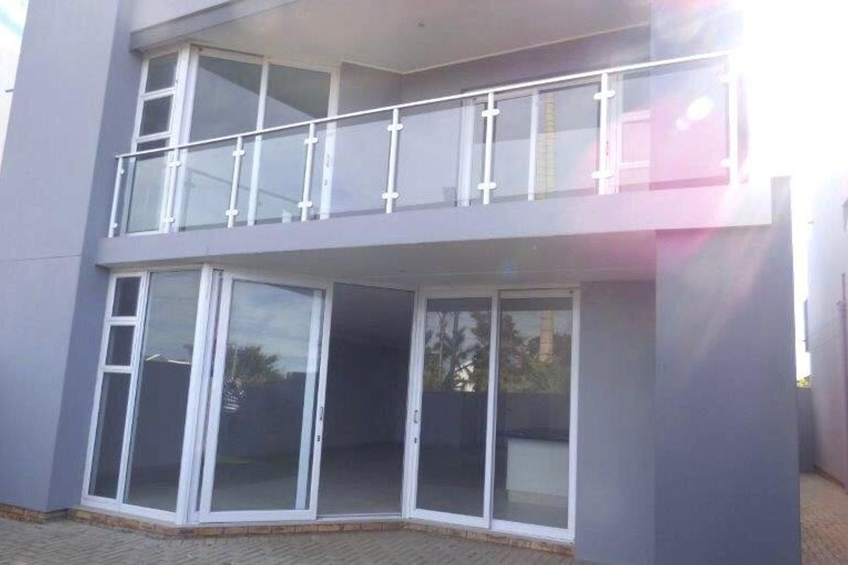 Rear view of a modern two-story house featuring a spacious upper balcony with glass balustrades and ground-level sliding doors opening onto a paved patio.
