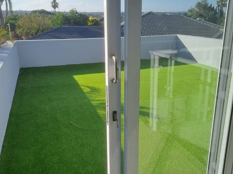 A private balcony featuring artificial turf flooring, enclosed by a parapet wall, accessible via a sliding glass door with views of the surrounding area.