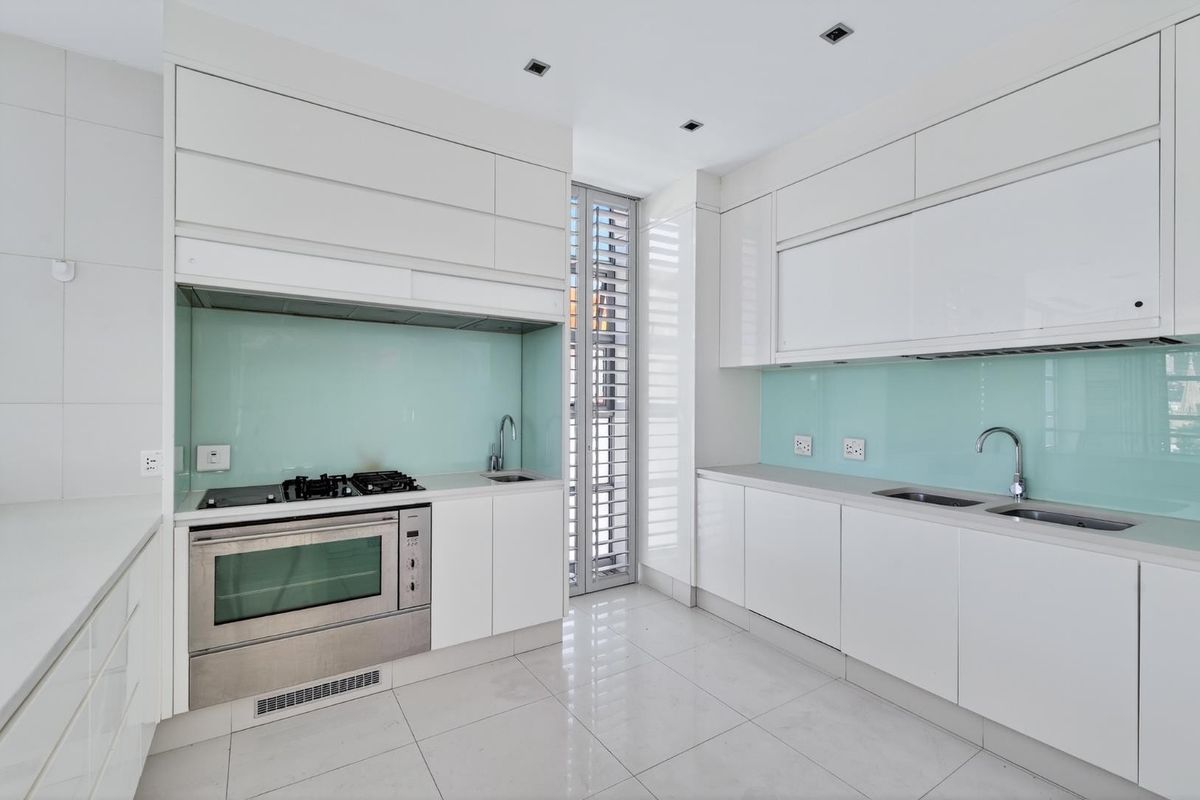Modern kitchen featuring sleek white cabinetry, integrated stainless steel oven, gas hob, and a light blue glass splashback with double sink.