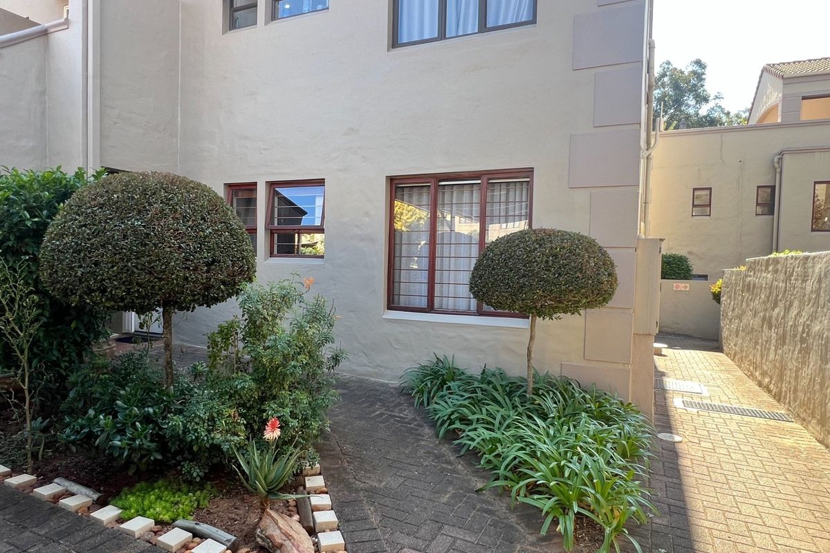 Exterior view of the apartment building featuring a paved walkway, landscaped garden beds with manicured shrubs, and secure window bars.