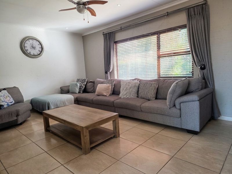 A comfortable living room featuring neutral tiled flooring, a ceiling fan, and large windows with fitted curtain tracks and blinds.