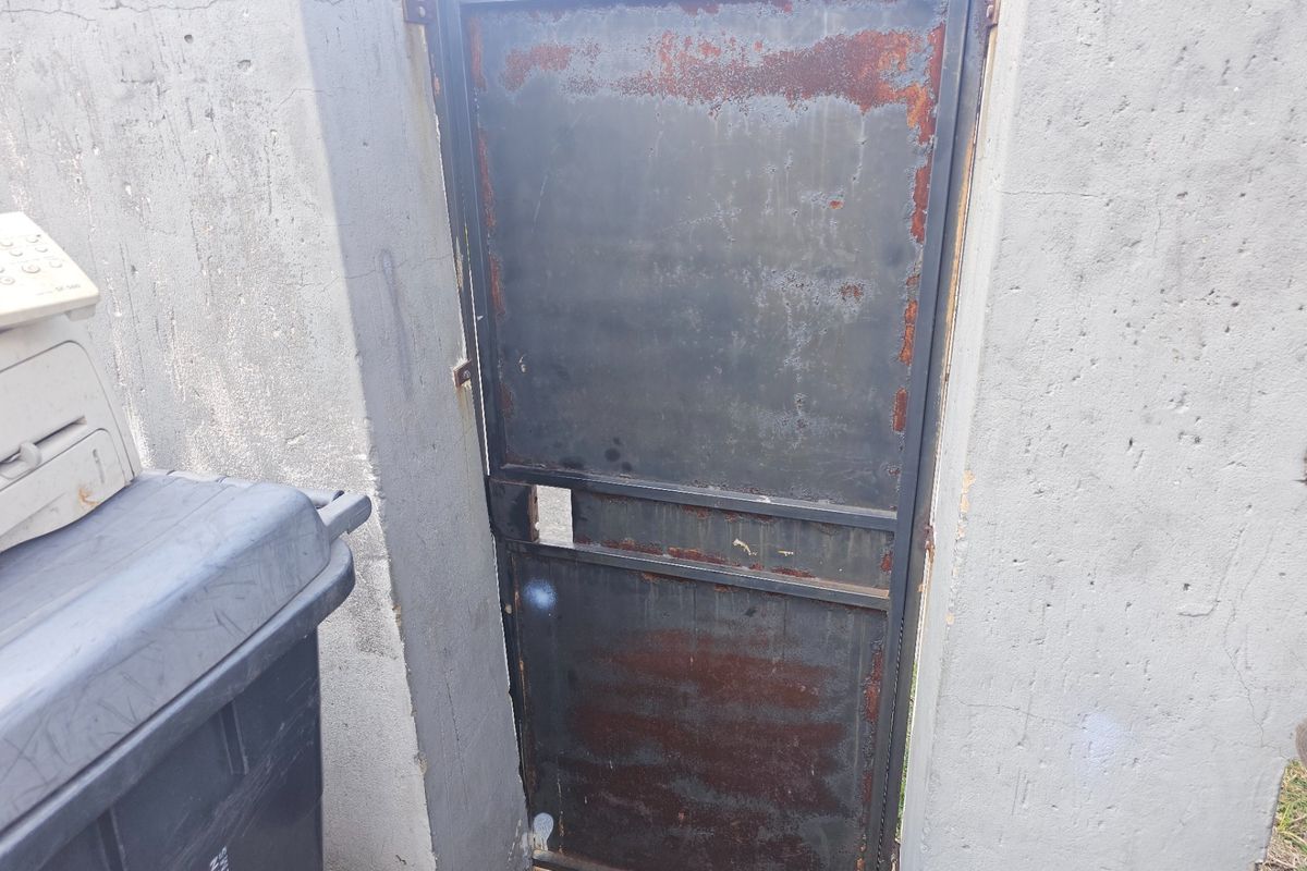 A weathered metal gate set within a concrete wall, showing significant signs of rust and structural deterioration.