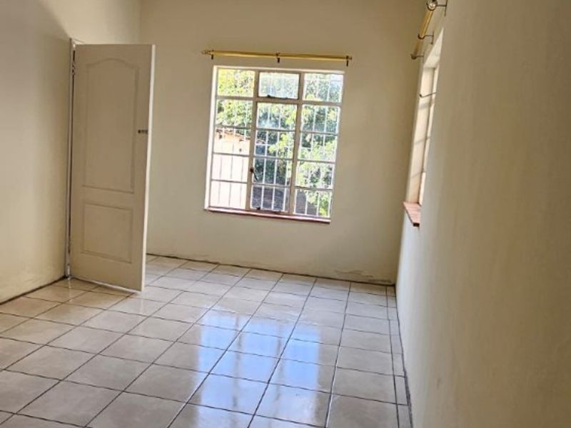 A bright, empty bedroom featuring neutral ceramic floor tiles, large windows for natural light, and a standard interior door.