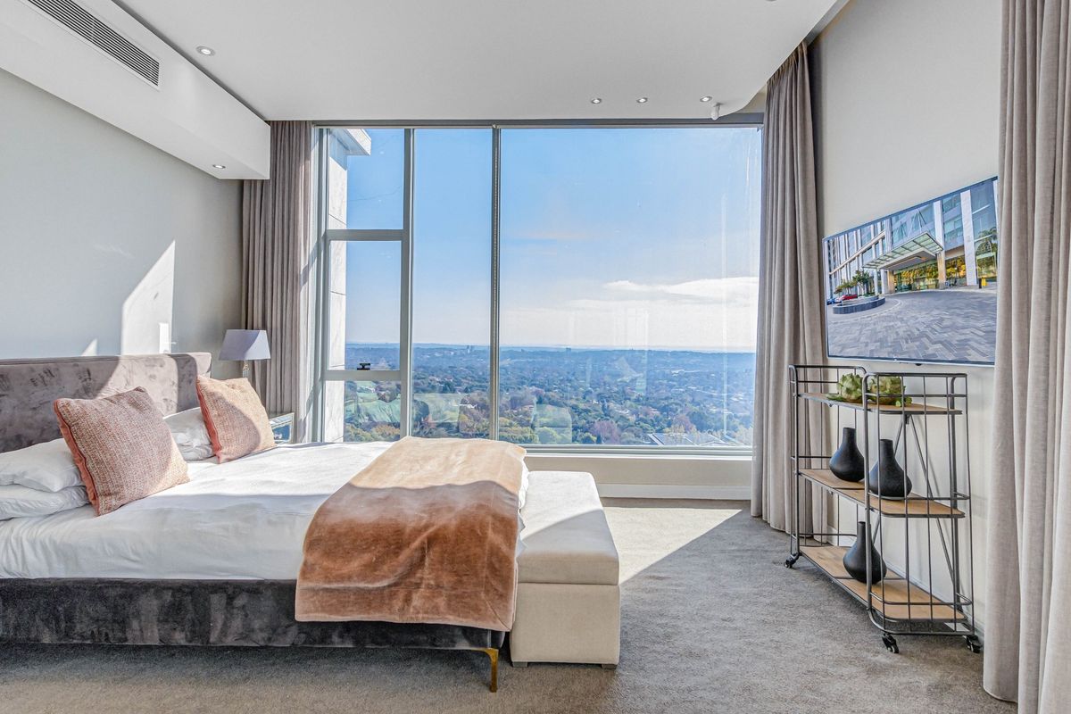 A bright, modern bedroom featuring floor-to-ceiling windows with expansive panoramic views, recessed ceiling lighting, and neutral carpeting.