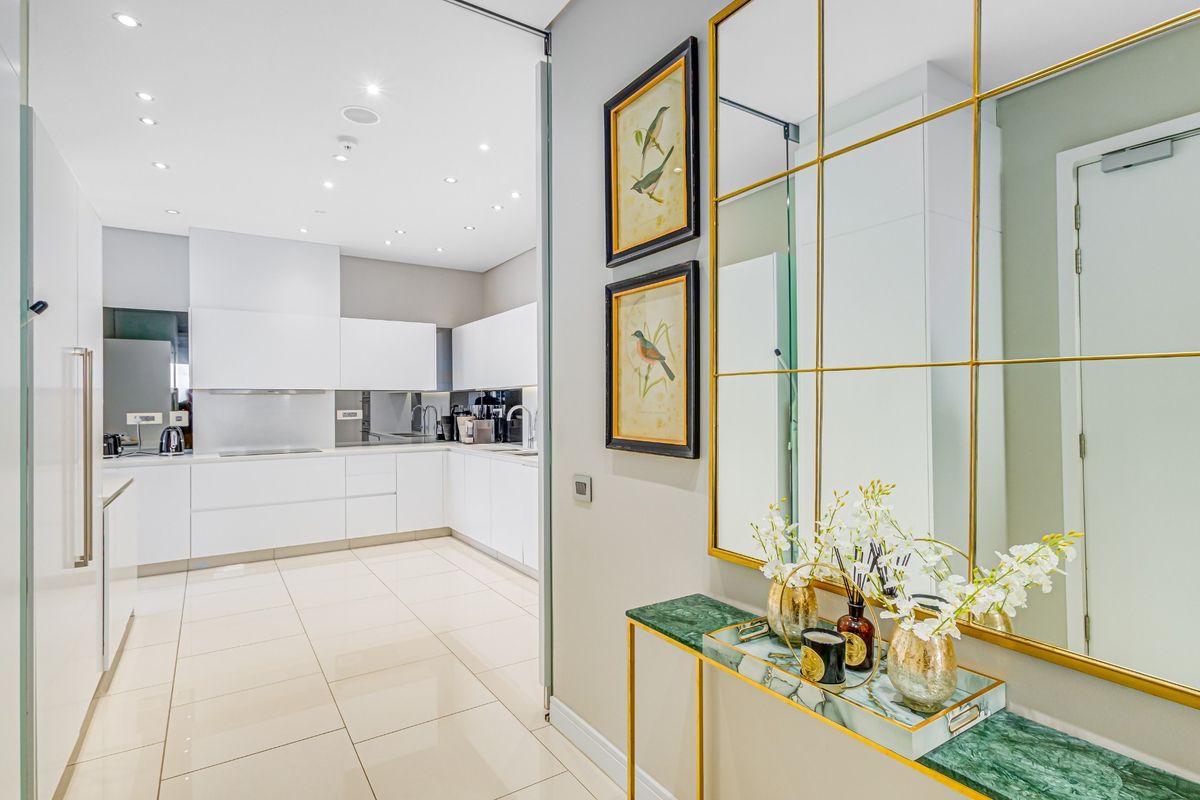 A modern, sleek kitchen featuring white cabinetry, integrated appliances, and polished tile flooring, viewed from an elegant entrance hallway with a large decorative mirror.