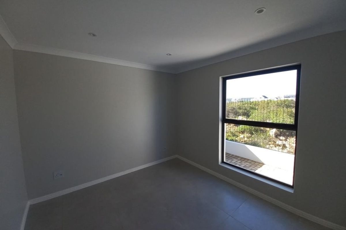 A bright, empty bedroom featuring neutral grey floor tiles, recessed ceiling lighting, and a large window with a view of the exterior.