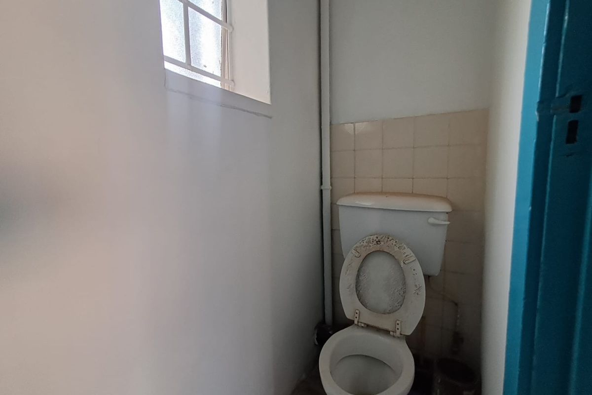 A small, dated bathroom featuring a standard toilet, tiled splashback, and a small window with security bars.