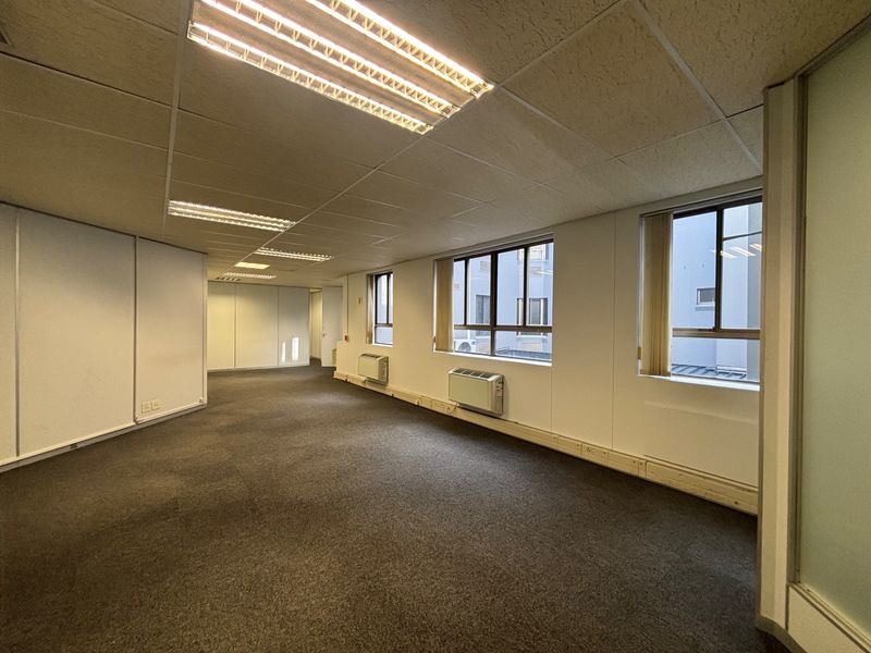 An empty, open-plan office space featuring suspended ceilings, fluorescent lighting, carpeted floors, and wall-mounted air conditioning units with natural light from multiple windows.