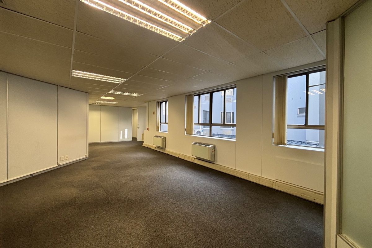 An empty, open-plan office space featuring suspended ceilings, fluorescent lighting, carpeted floors, and wall-mounted air conditioning units with natural light from multiple windows.