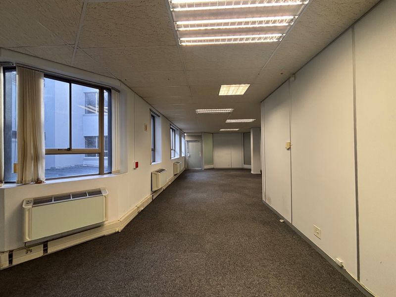 A spacious, empty office area featuring carpeted flooring, recessed ceiling lighting, and wall-mounted HVAC units, offering a functional workspace layout.