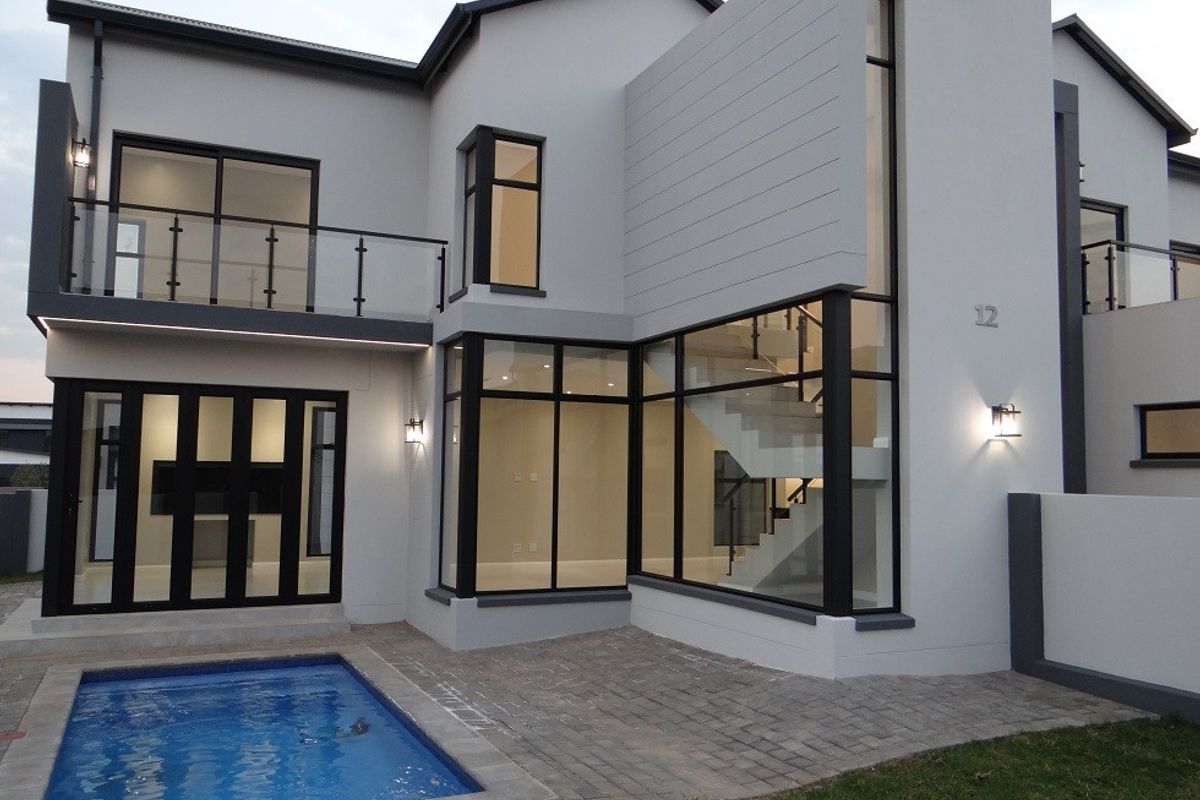 Modern double-story rear facade featuring large glass windows, a staircase, a glass-balustrade balcony, and a paved patio area with a swimming pool.