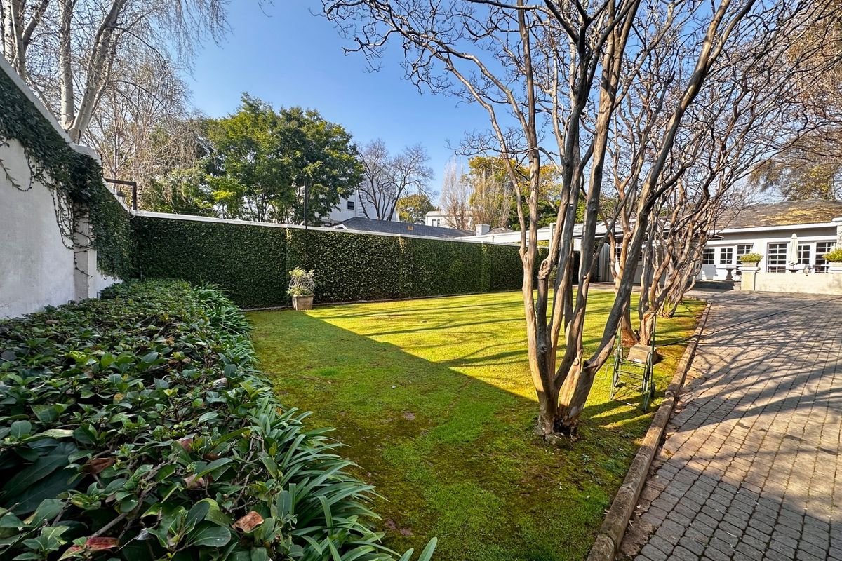 A well-maintained rear garden featuring a manicured lawn, mature trees, and a paved walkway, enclosed by high perimeter walls with established greenery.