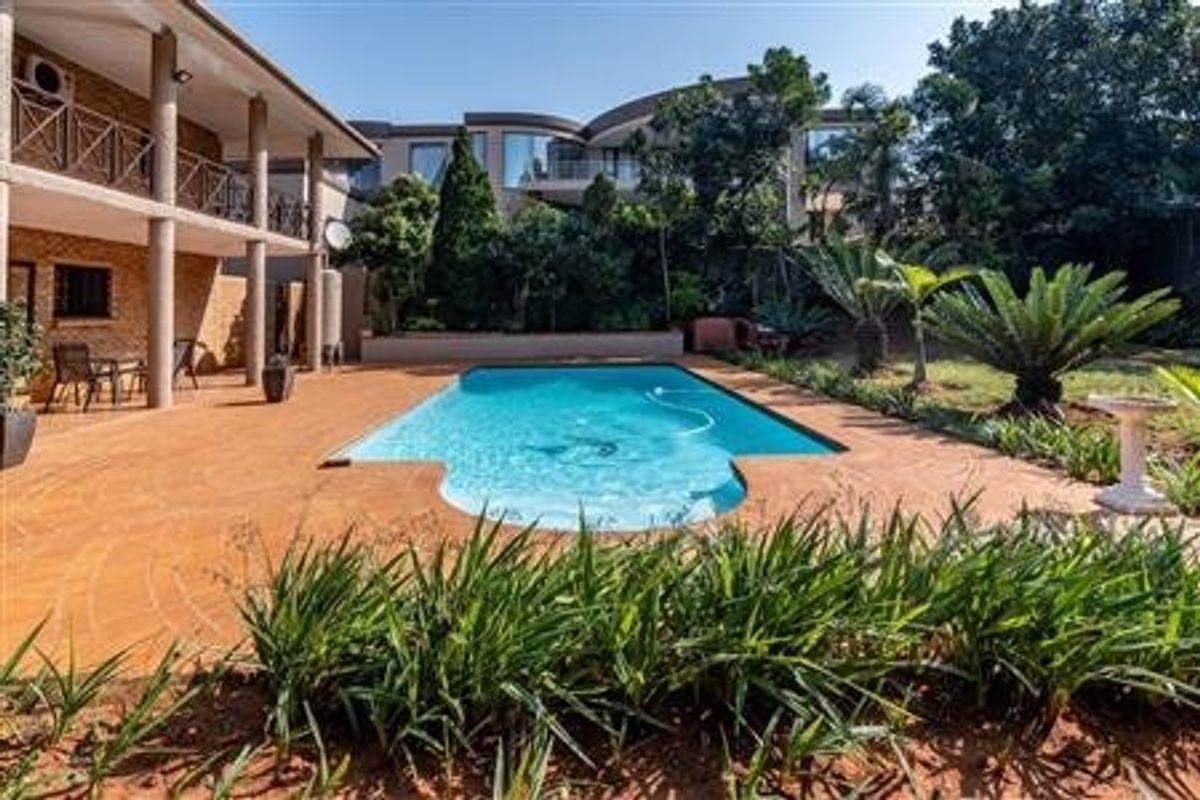 A spacious outdoor pool area featuring a paved patio, established garden landscaping, and a multi-level residential structure with balcony access.