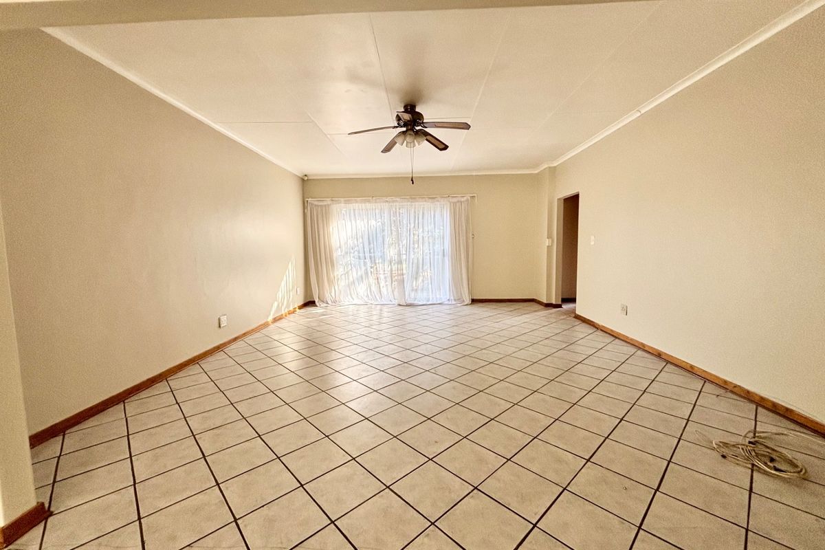 A spacious, empty living room featuring neutral tiled flooring, a ceiling fan, and a large sliding glass door providing natural light.