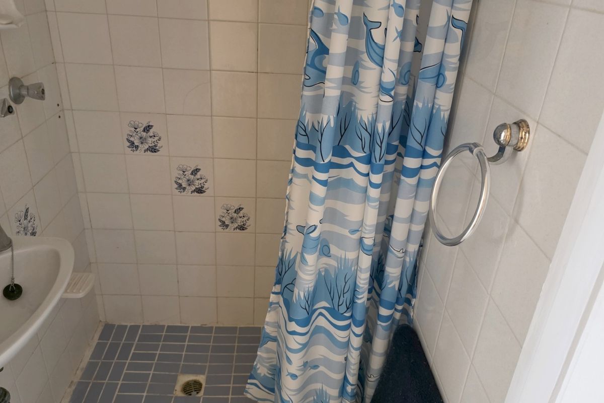 A compact bathroom featuring a shower area with a tiled floor and wall-mounted basin, showcasing dated finishes and fixtures.