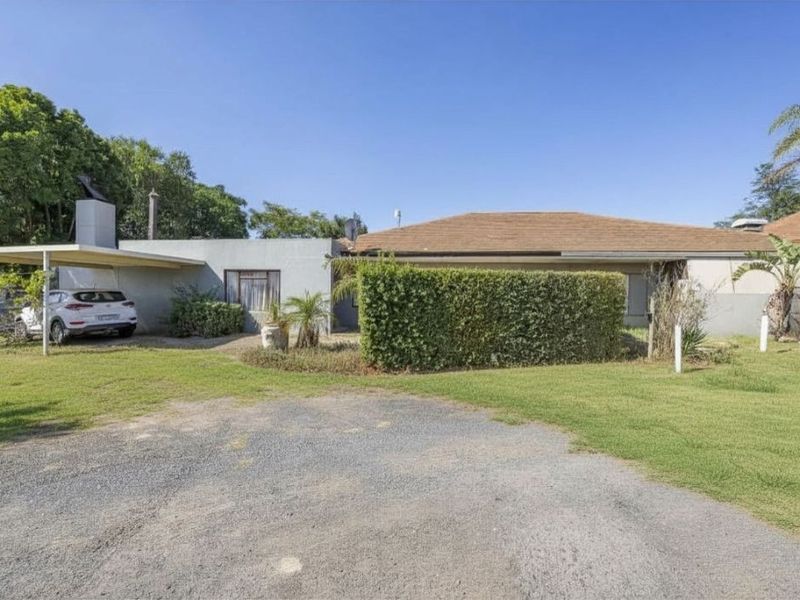 Front view of the farm property featuring a gravel driveway, carport, and established garden hedging.