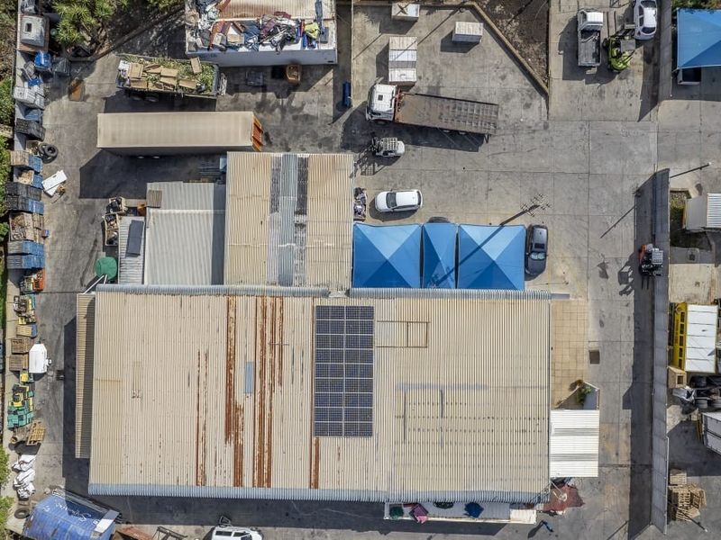 Aerial view of a farm property featuring a large industrial shed with solar panels, extensive concrete yard space, and various outdoor storage areas.