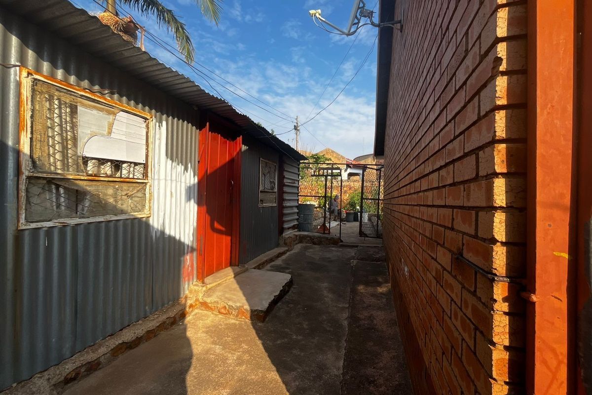 A narrow side passage between a corrugated iron structure and a brick building, featuring a concrete walkway and a metal gate.