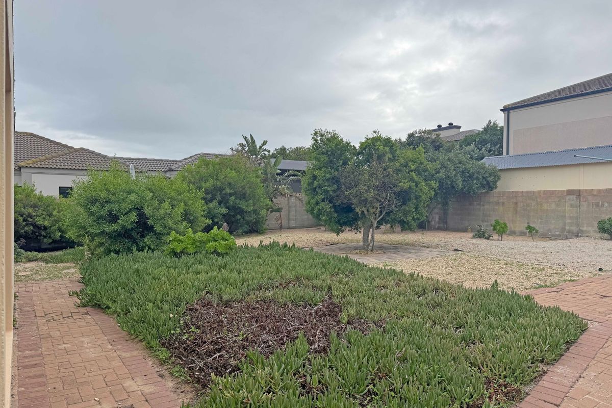 A spacious rear garden featuring paved pathways, established shrubs, and a large open area enclosed by concrete block boundary walls.