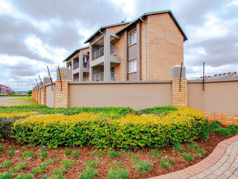 Exterior view of a modern brick apartment complex featuring perimeter walling with electric fencing, landscaped garden beds, and a paved walkway.