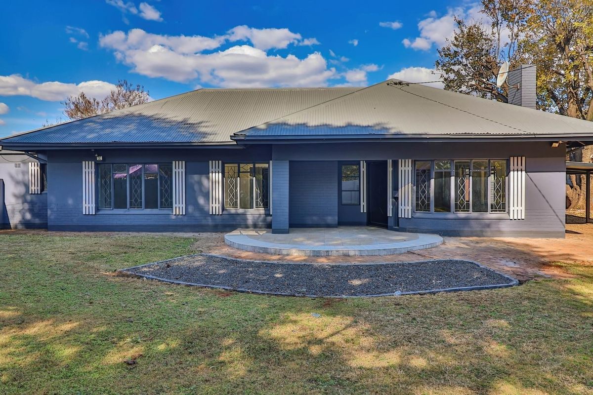 Rear view of a single-story house featuring a corrugated metal roof, painted brick exterior, leaded glass windows, and a paved patio area.