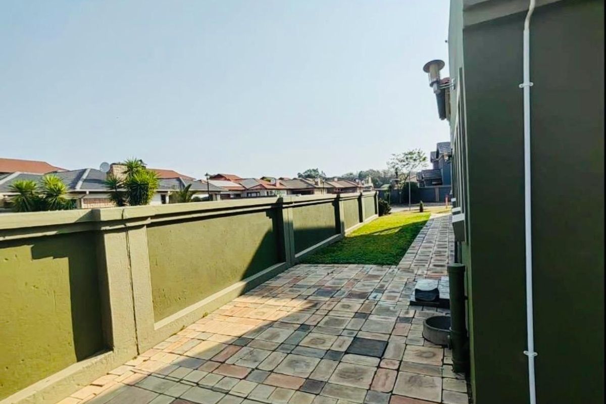A side exterior walkway featuring paved flooring, a boundary wall, and a small strip of lawn along the side of the house.