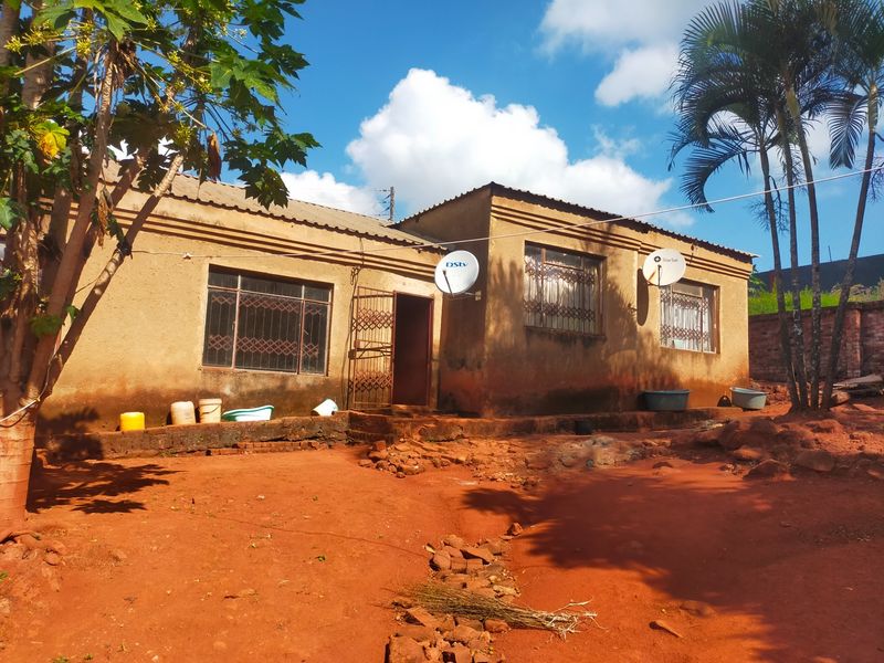 Single-story residential house exterior featuring a simple facade, metal security gates, and satellite dishes, set on an unpaved, natural earth plot.