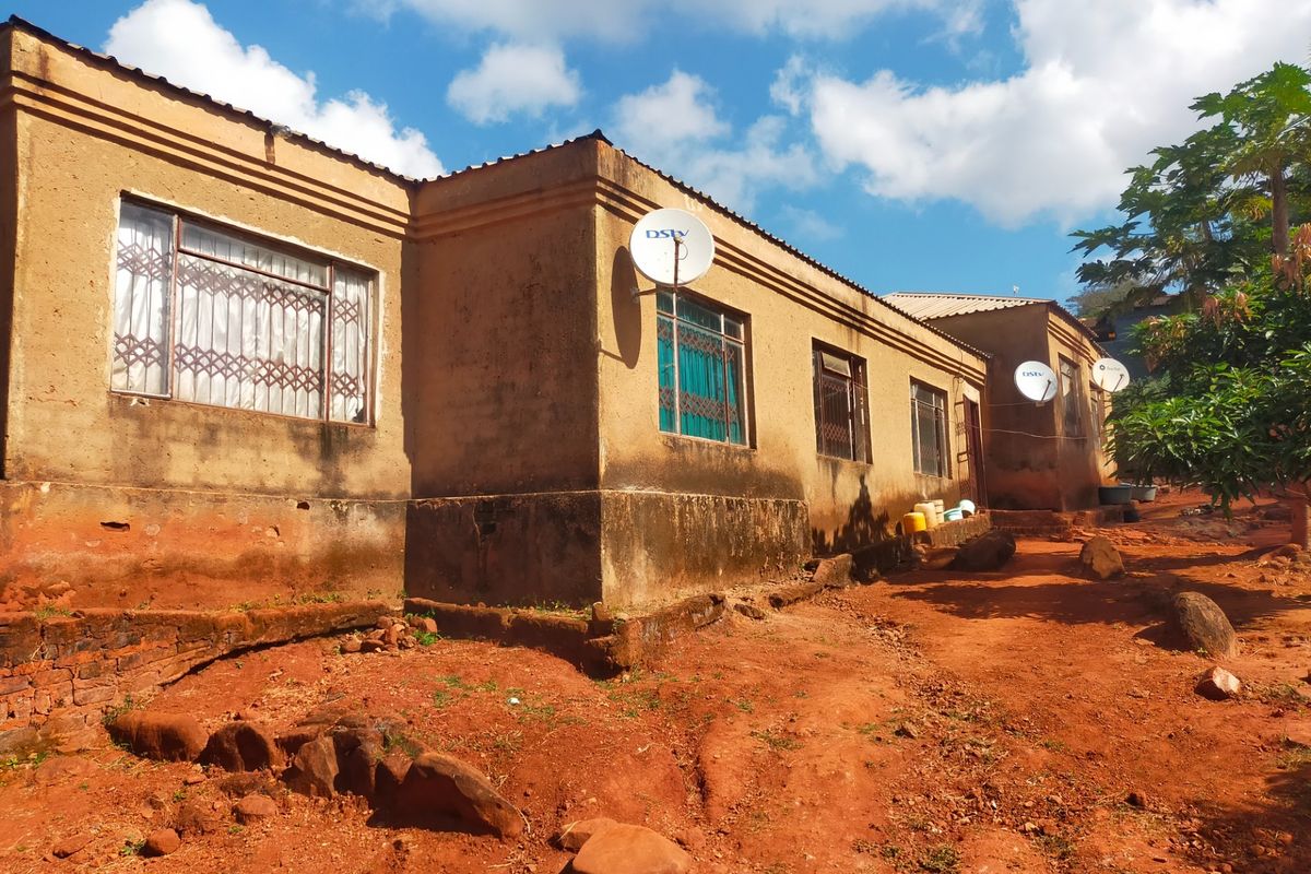 Exterior view of a single-story house featuring plastered walls, metal security window grilles, and satellite dishes, set on an unpaved, uneven plot.