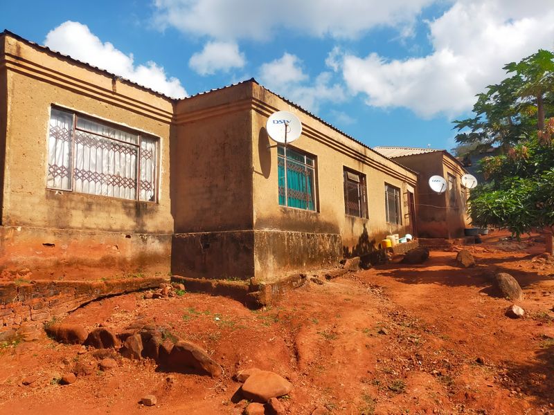Exterior view of a single-story house featuring plastered walls, metal security window grilles, and satellite dishes, set on an unpaved, uneven plot.