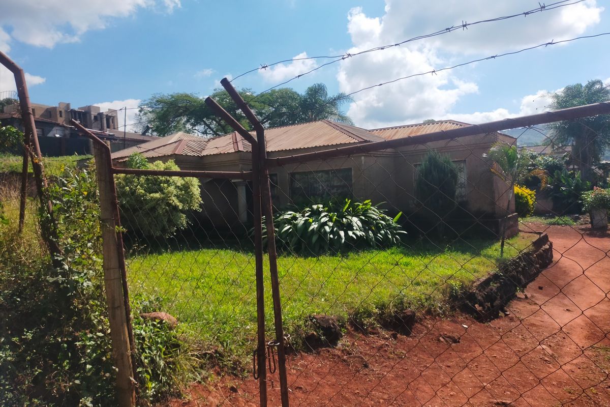 Front view of a single-story house featuring a corrugated metal roof, set behind a chain-link fence with an overgrown front garden.