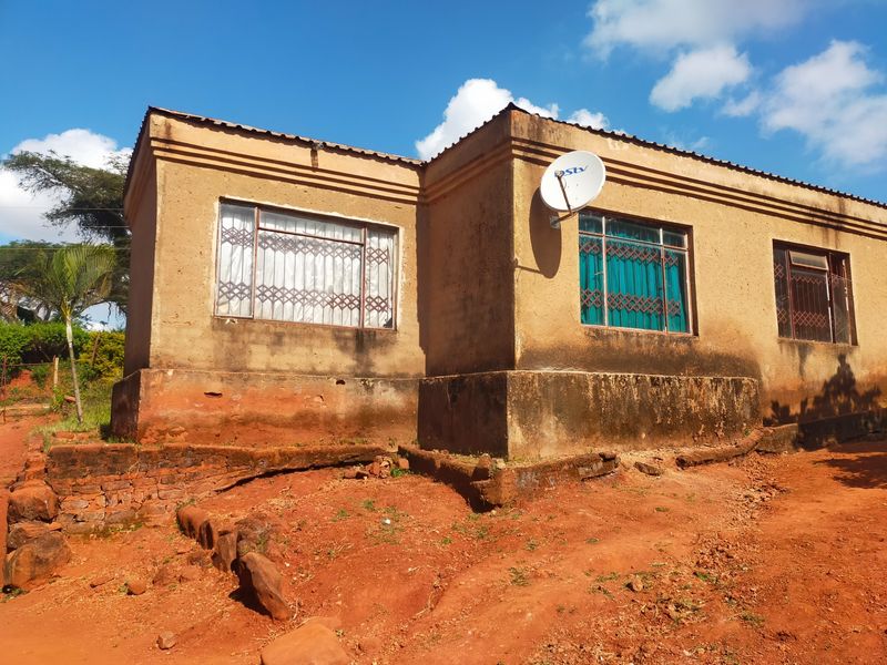Exterior view of a single-story house featuring metal security window grilles and a satellite dish, showing significant wear to the exterior walls and foundation.