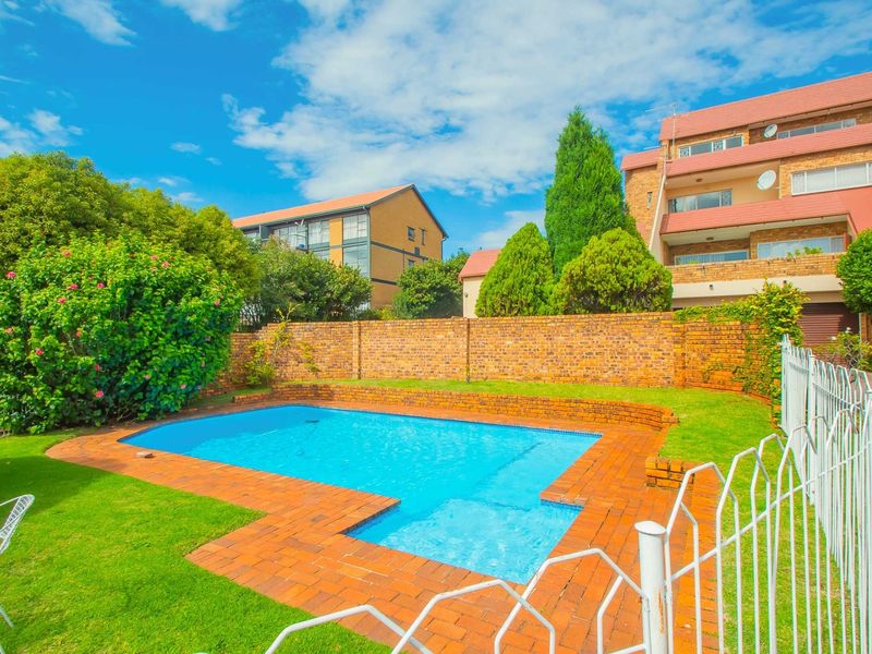 A well-maintained communal swimming pool area featuring brick paving, established garden landscaping, and secure metal perimeter fencing within an apartment complex.