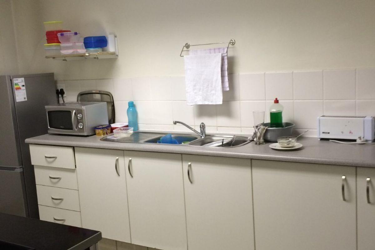 A functional kitchenette area featuring white cabinetry, a stainless steel sink, tiled splashback, and tiled flooring within an industrial workspace.