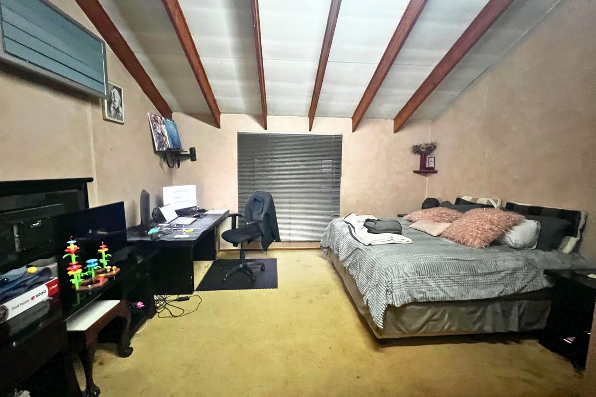 Spacious bedroom featuring exposed timber ceiling beams, carpeted flooring, and a large window with blinds, offering a functional layout for sleeping and workspace.